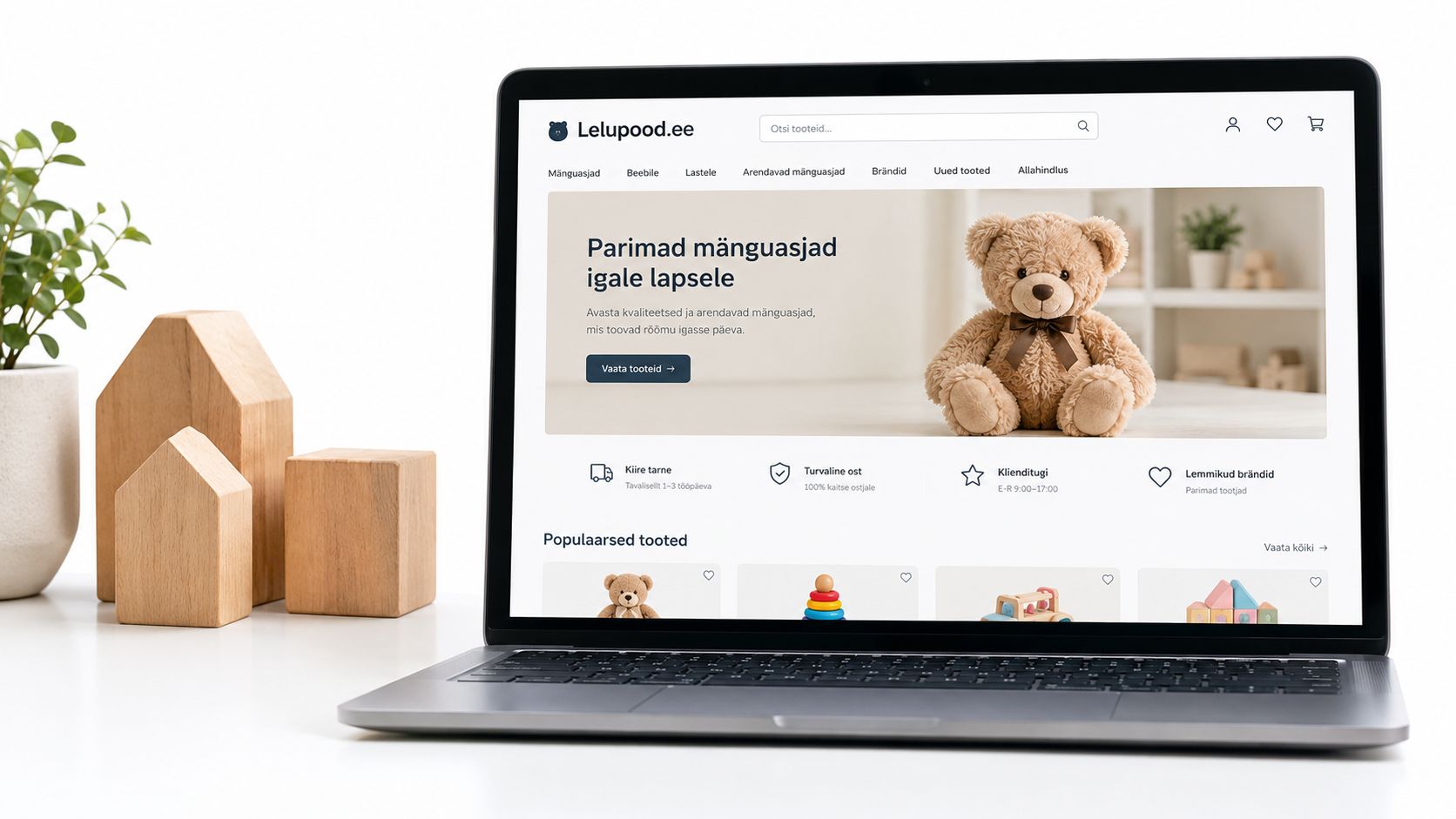 Sülearvuti ekraanil avatud mänguasjapood Lelupood.ee, mille kõrval asuvad puidust majakujulised dekoratsioonid valgel laual.